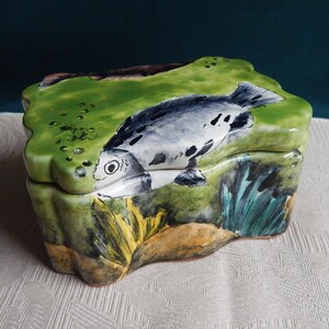May include: A ceramic box with a wavy, green lid featuring a hand-painted fish with black and white markings. The box is decorated with green, yellow, and blue paint, depicting an underwater scene.