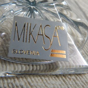 May include: A close-up of a white label with gold lettering that reads "Mikasa&reg; Slovenia". The label is attached to a clear glass object.
