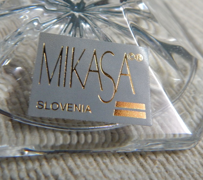 May include: A close-up of a white label with gold lettering that reads "Mikasa&reg; Slovenia". The label is attached to a clear glass object.
