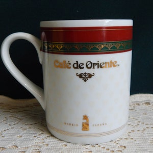 May include: White ceramic mug with a curved handle. The mug features the text "Café de Oriente" in gold lettering, along with decorative gold accents and a red band at the top. The bottom of the mug reads "Madrid España."