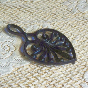 May include: A black cast iron trivet shaped like a leaf with an openwork design featuring swirling, curved lines and small circular accents. The object includes a circular handle at the top, providing a functional grip for this kitchen accessory.