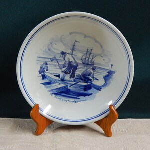 May include: A blue and white ceramic plate with a scene of people working on a boat. The plate has a blue rim and the words "De Scheepstimmerman" are written on the bottom.