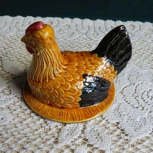 May include: A ceramic hen-shaped dish with a yellow body, black tail feathers, and a red comb. The dish is designed to hold eggs and is sitting on a round, textured base. The hen is a decorative kitchen item.