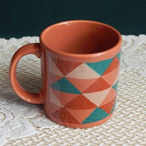 May include: A ceramic mug with a terracotta-colored exterior and handle. The mug features a geometric pattern of teal, peach, and orange triangles. The mug is sitting on a white lace tablecloth.