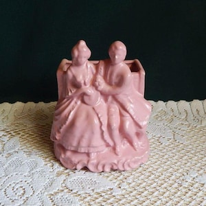 May include: A pink ceramic planter featuring a couple in period clothing. The figures are sculpted in relief, with the woman holding a musical instrument. The planter has a rectangular shape and is painted in a solid pink color.