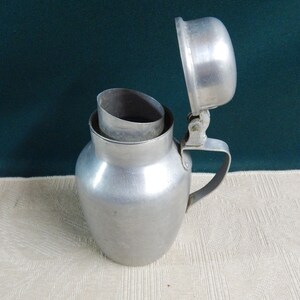 May include: A silver aluminum pitcher with a hinged lid. The pitcher has a rounded body and a handle. The lid is open and the pitcher is sitting on a white surface.