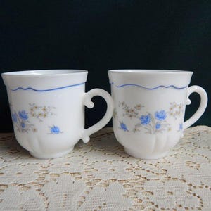 May include: Two white ceramic coffee mugs with blue and brown floral designs. The mugs have a scalloped rim and a blue trim.