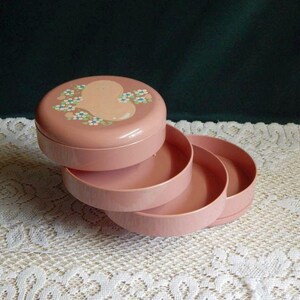 May include: A pink tiered jewelry box with a floral design on the top lid. The box has three tiers and is made of plastic.