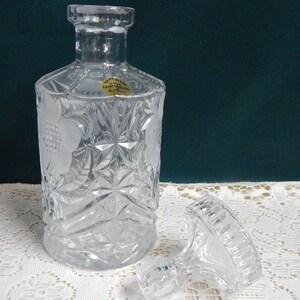 May include: A clear glass decanter with a textured design and a removable stopper. The decanter has a label that reads "Hand Made Lead Crystal".