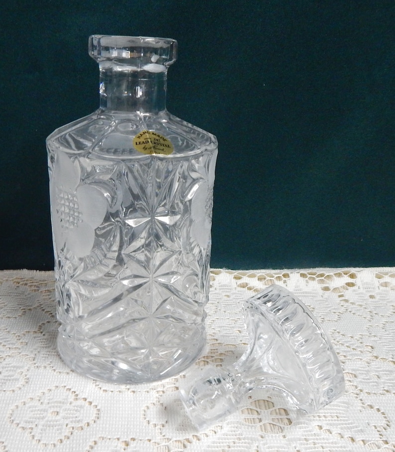 May include: A clear glass decanter with a textured design and a removable stopper. The decanter has a label that reads "Hand Made Lead Crystal".