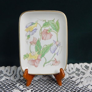 May include: A white porcelain trinket dish with a gold rim and a floral design featuring pink, yellow, blue, and green flowers. The dish is on a wooden stand.