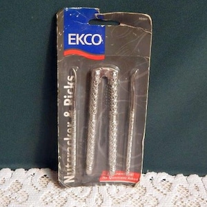 May include: A silver-toned nutcracker and picks set in a clear plastic package. The package has a blue and red EKCO logo. The set includes a nutcracker and two picks. The text "Nutcracker & Picks" is printed on the package.