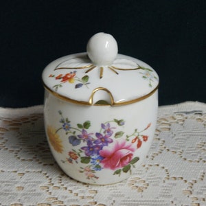 May include: A white porcelain honey pot with a floral design and a gold trim. The lid has a small spout for pouring.