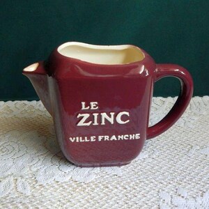 May include: A burgundy ceramic pitcher with a handle and spout. The pitcher has the words "LE ZINC VILLE FRANCHE" in white lettering. The interior is a cream color.