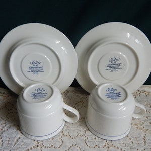 May include: Set of two white Lenox Chinastone teacups and saucers with a blue trim. The bottom of the cups and saucers have the Lenox Chinastone logo in blue.