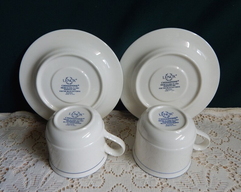 May include: Set of two white Lenox Chinastone teacups and saucers with a blue trim. The bottom of the cups and saucers have the Lenox Chinastone logo in blue.