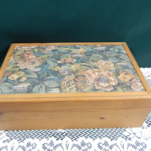 May include: A rectangular wooden box with a floral tapestry lid. The tapestry features roses and other flowers in shades of pink, yellow, and green against a blue background. The box is made of light-colored wood.