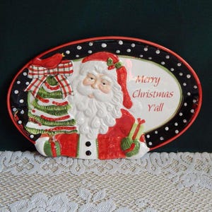 May include: Oval ceramic Christmas platter featuring Santa Claus with a red hat and suit, holding a green and red Christmas tree. The platter has a black border with white polka dots and the text "Merry Christmas Y'all".