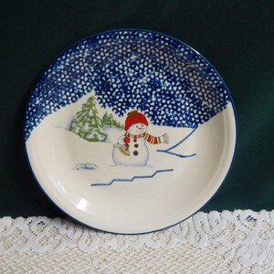 May include: A white ceramic plate with a blue dotted border and a winter scene. The plate features a snowman wearing a red hat and scarf, holding an axe. A snowy landscape with trees is also depicted.