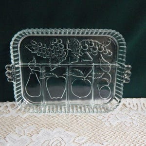 May include: Clear glass serving tray with a rectangular shape and a scalloped edge. The tray features embossed fruit designs, including grapes, pears, and apples. It has two small handles on either side, perfect for serving appetizers or desserts.