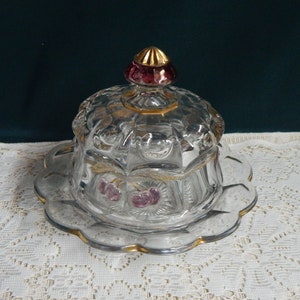 May include: A clear glass butter dish with a gold-trimmed lid and a pink glass knob. The dish is decorated with a pattern of cherries and gold accents.