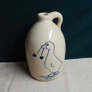 May include: A white ceramic pitcher with a blue and white hand-painted design of a goose holding two hearts in its beak.