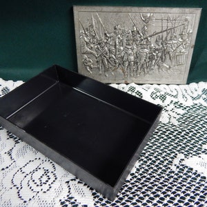 May include: A silver metal box with a raised image of a group of people in military attire. The box is open and the inside is black.