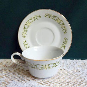 May include: White teacup and saucer set with a gold rim and green floral design. The teacup has a curved handle and the saucer is round. The set is placed on a white lace tablecloth.