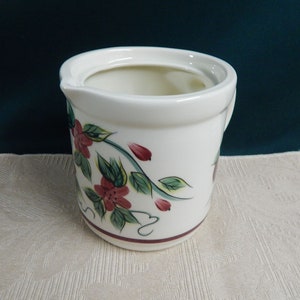 May include: White ceramic pitcher with a floral design featuring pink flowers, green leaves, and red accents. The pitcher has a spout and a handle.