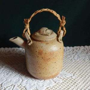 May include: A small, speckled ceramic teapot with a woven handle. The teapot is a light tan color with darker brown spots and a matching lid. The handle is made of a twisted, natural-colored material. The teapot is sitting on a white lace tablecloth.