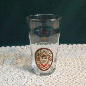 May include: A clear pint glass with a circular George Killian's Irish Red label. The label features a horse, the words "Premium Lager", and "Irish Red". The glass is empty and sits on a white lace tablecloth.