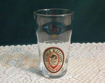 May include: A clear pint glass with a circular George Killian's Irish Red label. The label features a horse, the words "Premium Lager", and "Irish Red". The glass is empty and sits on a white lace tablecloth.