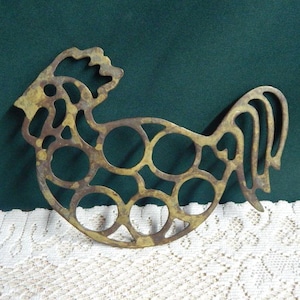 May include: A vintage brass rooster-shaped trivet with a cutout design. The trivet has a detailed comb and tail, and a series of circular cutouts within the body. The metal has a tarnished, aged appearance, suggesting it is an antique or vintage piece.