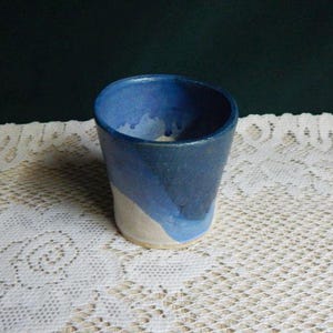 May include: A handmade ceramic cup with a gradient of blue hues. The cup transitions from a light beige at the base to shades of blue at the top. The interior is a darker blue. The cup is sitting on a white lace tablecloth.