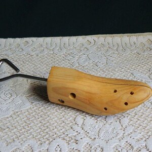 May include: A light-colored wooden shoe tree featuring a smooth, polished finish with visible natural wood grain. The device includes a metal rod extending from the heel area, ending in a triangular handle for manual adjustment and handling.
