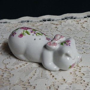 May include: A white ceramic pig figurine with pink flowers and a pink bow. The pig is lying on its side with its eyes closed.