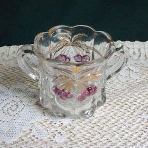 May include: A clear glass sugar bowl with two handles and a scalloped rim. The bowl is decorated with gold-colored leaves and purple cherries. The bowl is sitting on a white lace tablecloth.