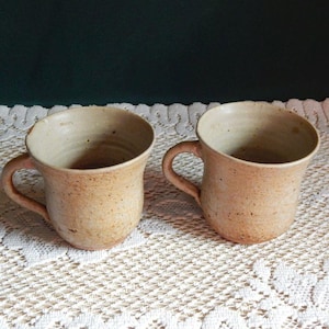 May include: Two handmade ceramic mugs with a speckled, tan glaze. Each mug has a rounded body, a curved handle, and a slightly flared rim. The mugs are displayed on a white lace tablecloth, suggesting a rustic or farmhouse aesthetic.