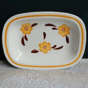 May include: White rectangular serving dish with a yellow and brown floral design. The dish has a yellow border and is decorated with three yellow flowers and brown leaf accents. The dish is on a white lace tablecloth.