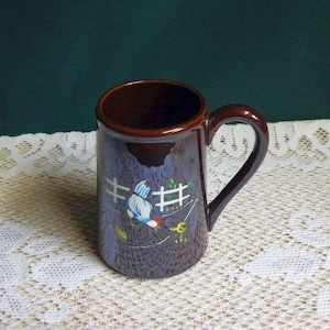 May include: A brown ceramic mug with a handle. The mug has a blue background with a white fence and a bird design. The interior and handle are a darker brown color. The mug is sitting on a white lace tablecloth.
