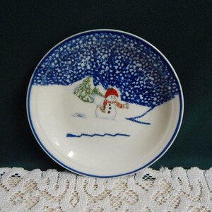 May include: White ceramic plate with a blue rim and a winter scene. The plate features a snowman wearing a red hat and scarf, with a snowy background and a blue sky. A small green tree is also visible.