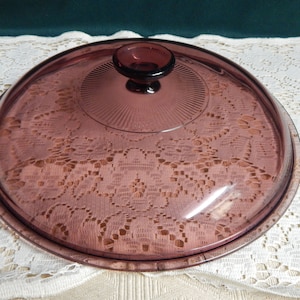 May include: A round, translucent purple glass lid featuring a central knob handle and a radiating sunburst pattern around the center. The surface of the lid is smooth, and the glass has a deep plum hue that allows light to pass through it.