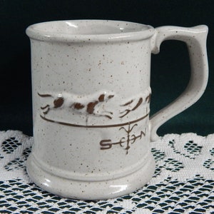 May include: White ceramic mug with a brown speckled glaze. The mug features a brown silhouette of a running dog and a compass design with the letters "S", "E", "W", and "N".