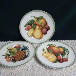 May include: Three white ceramic plates with fruit designs. Each plate features a different arrangement of colorful fruits, including pears, plums, cherries, and a pineapple, with green leaves. The plates have a green border.