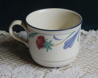 May include: Cream-colored teacup with a blue rim and handle. The cup is decorated with a floral pattern featuring red, blue, and green leaves and flowers. The cup is sitting on a white lace tablecloth.