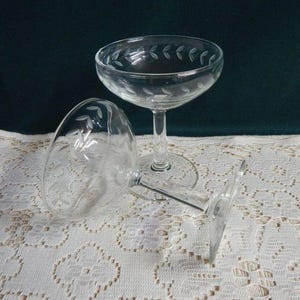 May include: Two clear glass champagne coupes with a delicate etched leaf design. The glasses are on a white lace tablecloth.