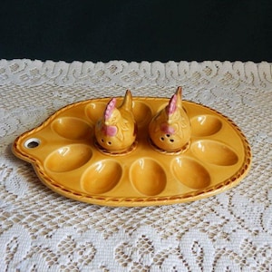 May include: A yellow ceramic escargot dish with 12 indentations, accompanied by two chicken-shaped salt and pepper shakers. The shakers are yellow with pink accents. The dish has a handle and a decorative border.