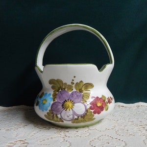May include: A white ceramic basket with a green handle and floral design. The flowers are painted in shades of purple, blue, red, and green.