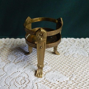 May include: A circular brass stand featuring three ornate legs with claw-style feet and a decorative band around the center. The metal has a warm, metallic gold tone with intricate embossed patterns along the rim and the vertical support structures.