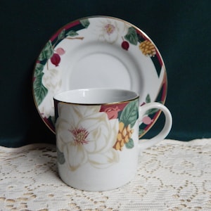 May include: A white teacup and saucer with a floral design featuring white magnolia flowers, green leaves, and red berries. The cup has a gold rim and the saucer has a gold rim and a burgundy band.